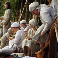 A group of individuals dressed in traditional, medieval-style clothing sits and stands near a wooden structure with greenery at the Semigallian festival. Some are engaged in crafts such as embroidery and braiding. Most are wearing light-colored garments and head coverings, creating a historical and communal atmosphere.