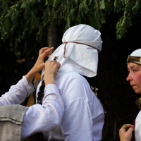 Three people dressed in historical white garments and head coverings. One person adjusts the back of another's headwear adorned with gold decorations. The third person watches. They are outdoors underneath tree branches, likely participating in a 2009 Semigallian Days cultural event or reenactment.