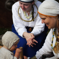 Two women in traditional clothing and headscarves sit on the ground with a baby, who is also dressed in traditional attire. The woman on the right smiles at the baby, while the other woman looks on. Their close interaction suggests they are part of a family or community gathering at a Semigallian festival.