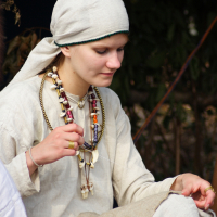 A person dressed in historical attire, wearing a beige headscarf and matching tunic, sits with a focused expression at the Semigallian Days festival. They have multiple necklaces with various beads and ornaments. Their right hand rests on their lap, holding part of their garment, while the background is blurred with greenery.