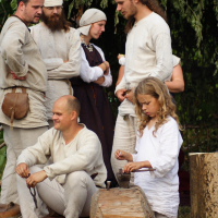 A group in historical attire gathers outdoors during Semigallian Days 2009. A seated child hammers metal on a wooden block while two men, one bald and the other with long hair, watch. Others, including a bearded man in a white cap, engage in conversation amidst leafy branches and logs in the background.
