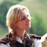 A woman with blonde hair braided on one side is dressed in historical attire, including a dark shawl, a large ornate brooch, and a twisted necklace. She is outdoors at the Semigallian festival, with blurred greenery in the background, and gazes thoughtfully to her right, bathed in soft natural light.