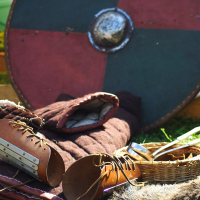 A medieval setup with a round shield featuring quadrants in red and green, leather armor and bracers, and a basket with metal and wooden utensils. On the ground are a fur rug, a folded blanket, and additional leather gear, all lit by sunlight—an homage to Semigallian culture celebrated during the 2009 event.