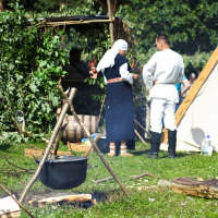 A historical reenactment scene set outdoors. In the foreground, a black cauldron hangs over a fire tripod with logs nearby. Two people in period clothing, one in a dark dress with a white headscarf and the other in a white tunic and black boots, stand conversing near a green bush during Semigallian Days 2009.