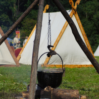 A tripod made of sturdy wooden poles supports a hanging cauldron over a small fire. The cauldron is suspended by a chain and sits above a partially burned log. In the background, white canvas tents with wooden frames are visible amidst green foliage, evoking the rustic charm of the 2009 Semigallian festival.