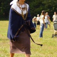 A woman walks on grass, wearing a traditional outfit: a white headscarf, a shawl with gold accents draped over her shoulders, a white top, a brown skirt, and tan shoes. She wears multiple necklaces and holds a strap. The scene is set during Semigallian Days in 2009. People and trees are visible in the background.