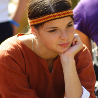 A young woman, dressed in a brown medieval-style garment with white cuffs, rests her chin on her hand. She wears a colorful woven headband across her forehead. Her expression appears thoughtful and serene as sunlight illuminates her face. Blurred people are in the background at the Semigallian festival.