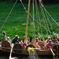A group of people are rowing a wooden boat designed like a Viking ship during Semigallian Days '09. Equipped with a central mast and round shields hanging on the sides, the boat moves along a calm waterway surrounded by lush greenery. The group, dressed in historical attire, rows as water splashes near the boat.