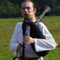 A person with long hair tied back, dressed in a white medieval-style tunic, plays a black bagpipe outdoors during the Semigallian festival. The individual is in a grassy field with dense green foliage in the background. Wearing a belt with a pouch attached to it, they have a focused expression while playing.