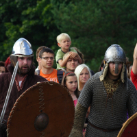 Two people dressed in medieval warrior costumes with helmets, chainmail, and round shields stand in the foreground. They hold swords and appear engaged in an activity. In the background, a diverse crowd, including a man holding a child on his shoulders, watches attentively. Trees are visible behind the crowd at Semigallian Days 2009.