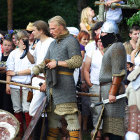 A group of people, some in medieval attire, participate in an outdoor event. In the center, two men dressed as Semigallian warriors in chainmail and leather armor stand in conversation. One holds a sword, and a crowd of onlookers, including children, is gathered behind them. Trees fill the background.