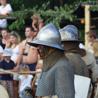 Two medieval reenactors dressed in chain mail and metal helmets stand side by side, holding swords and shields at the Semigallian Days event. They are part of a historical reenactment, watched by a crowd of onlookers in modern clothing. Greenery and wooden structures decorate the background.