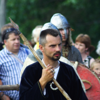 A man dressed in medieval clothing holds a spear, standing among a crowd at Semigallian Days 2009. He wears a dark tunic and has short hair and a beard. Behind him, a man in chainmail armor and a helmet is visible, along with other onlookers, some of whom are dressed in modern clothing. Trees are in the background.