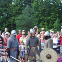 Three people dressed in medieval armor, including chainmail and helmets, participate in the 2009 Semigallian Days re-enactment event. The person on the right holds a round shield and a sword. The scene is outdoors, with a crowd of onlookers in summer clothing and green trees in the background.