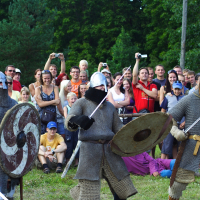 Three people dressed in medieval chainmail armor, helmets, and holding swords and shields, engage in a reenactment during Semigallian Days 09. A large, cheering crowd of men, women, and children look on and take photos. Trees and greenery form the backdrop of this outdoor event.