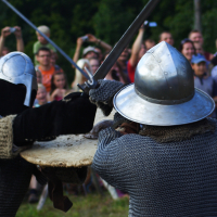 During the Semigallian Days '09, two knights in chainmail and metal helmets engage in sword combat during a reenactment. Both have raised swords clashing together and one carries a large shield. Onlookers in the background, some taking photos, watch the scene set outdoors among trees and greenery.