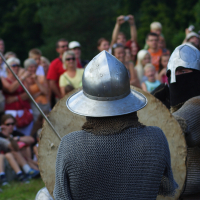 During the 2009 Semigallian Days, two people dressed in medieval armor and chainmail engage in a mock battle, holding swords and shields. They are surrounded by a crowd of spectators in casual attire, eagerly watching the performance, some taking photos. The event appears to be outdoors with trees in the background.