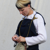 A person with short hair, half blonde, half black, wears a white long-sleeve shirt under a dark blue garment fastened with two golden brooches. They stand against a light-colored background, looking down while opening a small beige pouch hanging around their neck—reminiscent of the Semigallian days.
