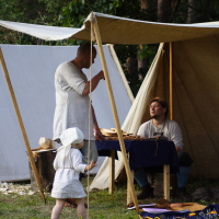 A historical reenactment scene from the 2009 Semigallian Days shows two adults and a child in period clothing under a canvas tent. One adult stands, facing another seated at a table with various wooden tools. The child, wearing a white bonnet and dress, appears to approach the table. The backdrop is a forested area.
