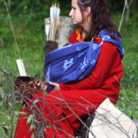 At the 2009 Semigallian Days, a woman in a red dress sits on a log outdoors, holding a mug in one hand and a baby wrapped in a blue cloth carrier in the other. The background features greenery, a wooden stump, and arrows in a quiver. She appears to be gazing into the distance.
