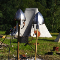 Two medieval helmets on wooden poles stand in the foreground of a historical reenactment campsite. In the background, there are canvas tents and trees, with a small campfire burning near the center. Various camp supplies are scattered around, suggesting an active campsite reminiscent of Semigallian Days 2009.