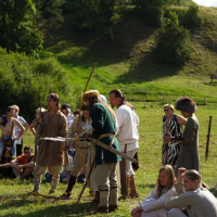 A group of people dressed in medieval clothing gather outdoors on a grassy field by a hillside during the Semigallian Days 2009. Some are standing with bows and arrows, while others are sitting on the grass. Behind them, spectators watch the scene. Trees and a small hill with a flag flying on top are in the background.