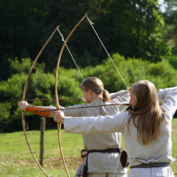 Two individuals dressed in Semigallian medieval-style clothing are outdoors, standing on a grassy area. They each hold traditional wooden bows, drawn back in an archery pose, ready to release arrows. A lush, green forest is visible in the background, with sunlight casting bright highlights.