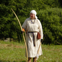 A person dressed in historical Semigallian medieval attire stands in a grassy field with a wooded background. They wear a checked tunic, a headscarf, and hold a longbow. A quiver with arrows is slung over one shoulder, and a brown pouch is attached to their belt. The scene is bright and green.