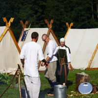 In an outdoor camp with white tents, three people in medieval attire stand by a campfire with a metal pot over it during the 2009 Semigallian Days. One person tends to the fire, while another, dressed in a green tunic and white headscarf, watches. Logs and a shield lie on the grass beside them. Dense trees are in the background.
