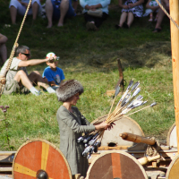 A person dressed in historical attire, wearing a fur hat, is holding a bundle of arrows while standing on a wooden Viking ship during Semigallian Days 2009. The ship is equipped with round shields painted in red and yellow stripes. Spectators, seated on grass in modern clothing, watch the scene from the background.
