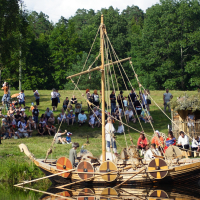 A group of people dressed in historical Viking costumes are gathered on a wooden longship with colorful shields attached to the sides. They are docked near the shore of a calm water body. On the grassy bank, a large crowd watches the scene, reminiscent of Semigallian Days 2009, with a thatched hut and dense trees in the background.