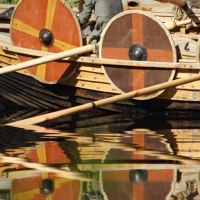 A person sits on the side of a Viking-style wooden boat, adorned with round shields featuring red and yellow patterns. Reflected in the calm water below, the scene captures the essence of the Semigallian festival. The background consists of grass and a wooden pole from the 2009 event.