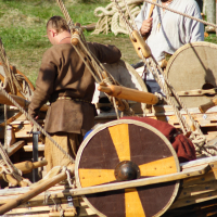At Semigallian Days 2009, two men dressed in historical clothing stand on a wooden boat. One man, facing away, wears a brown tunic while the other, in a white tunic with a black headband, looks forward holding a rope. Visible are circular wooden shields with yellow and brown patterns and various ropes across the boat.