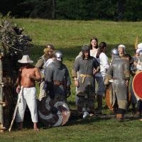 A group of people in medieval costumes gathers on a grassy field at Semigallian Days 2009. Some wear chainmail and helmets with swords and shields, while others are in light tunics. A shirtless man with a white hat stands near a wooden shelter covered with foliage. In the background, trees are visible.