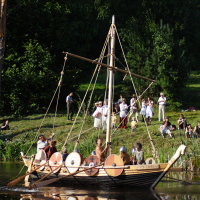 A group of people dressed in historical attire sail a Viking-style wooden boat with round shields on its sides during the Semigallian Days 2009 event. The boat glides on a calm river, reflecting the vessel and passengers. Spectators in varied dress watch from a grassy, tree-lined shore.