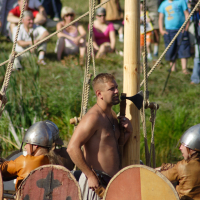 A shirtless man stands on a wooden Viking ship holding an axe during the 2009 Semigallian days. He is surrounded by three individuals in Viking helmets and armor, with round, painted shields adorning the ship's side. In the background, a crowd of people, seated and standing, observes the re-enactment.