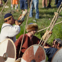 Three men dressed in historic Viking attire stand on a wooden ship adorned with round shields, some featuring crosses. One man blows into a horn, while another wears a fur hat and red tunic reminiscent of Semigallian warriors. A crowd in contemporary clothing watches from the background.