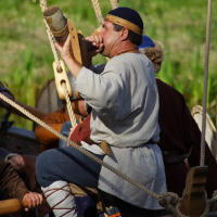 During Semigallian Days 2009, a person in historical attire, wearing a light grey tunic and a navy blue headband, blows into a horn while standing on a wooden boat. Several ropes are visible around the vessel, and other individuals in similar attire are partially seen against the lush greenery.