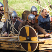 A group of people wearing Viking-style helmets and traditional clothing are on a wooden boat. The boat features oars, ropes, and a shield with a yellow and black design attached to the side. Behind them is a grassy area. The scene suggests a historical reenactment, possibly during Semigallian Days '09.