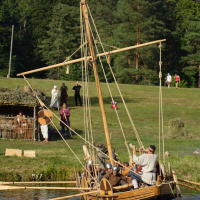 A wooden Viking ship with shields on the sides is floating on a body of water near a grassy shore during the Semigallian festival. People dressed in historical Viking attire, including helmets and tunics, are aboard the ship. In the background, there are trees and people in modern clothes observing the scene.