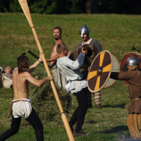 A group of people is engaged in a historical reenactment battle on grass during the Semigallian Days. They wear medieval costumes with tunics, helmets, and shields. Two of them clash with swords and shields, while another swings a large wooden paddle. In the background, a few others observe or rest on the grass.