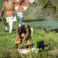 During the Semigallian Days event in 2009, a reenactment scene shows individuals in medieval attire and helmets engaged in a battle. One person stands over another who lies on the ground near a body of water, possibly tending or attacking. In the background, other participants wield shields and swords. The setting is grassy and outdoors.