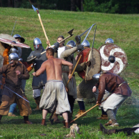 A group of reenactors are engaged in a mock medieval battle on a grassy field during the Semigallian Days festival. Some wear chainmail and helmets, while others are bare-chested. They wield wooden shields, axes, and swords. One fallen participant lies on the ground. The background features a hill and trees.