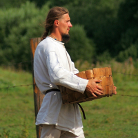 A man with long hair and a beard, dressed in a white linen shirt and pants with a black belt, walks through a grassy area carrying a wooden basket. The background features a wire fence and lush green trees and bushes, evoking the charm of the 2008 Semigallian Days. He seems to be in a rural or historical reenactment setting.