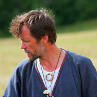 A bearded man with tousled hair, wearing a blue robe over a white shirt and a necklace with charms, stands against a blurred backdrop of greenery and open field. His head is turned to the right, looking downward, conveying a contemplative expression reminiscent of simpler Semigallian days.