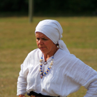 An elderly woman dressed in traditional attire stands outdoors at the 2008 Semigallian Days event. She wears a white headscarf and blouse, with a colorful beaded necklace around her neck. Her hands rest on her hips as she looks down thoughtfully, with a blurred background of grass and trees.