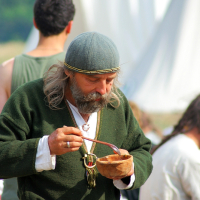 A bearded man in historical attire, including a green tunic and knitted cap, enjoys a meal from a wooden bowl using a spoon during Semigallian days. He stands in an outdoor setting with blurred figures and tents in the background, indicating a possible historical reenactment or festival.