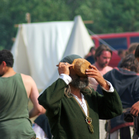 A person dressed in historical attire, including a green tunic and a belt with a pouch hanging from it, drinks from a wooden bowl at the 2008 Semigallian festival. Others in medieval-like costumes are in the background, with tents and greenery also visible, suggesting a historical reenactment or fair.