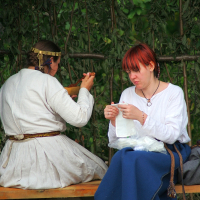 Two individuals in historical clothing sit outdoors on a wooden bench during the 2008 Semigallian Days festival. The person on the left, with braids and a headband, is eating from a bowl. The person on the right has red hair and is focused on sewing white fabric. They are under a leafy canopy, with a bag resting beside them.