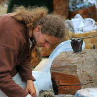 A person with long, curly hair, wearing a brown garment, is sitting on the ground and working with tools and materials, possibly engaging in a craft or historical reenactment. They are surrounded by wooden items, fabric, and other crafting materials at what appears to be the Semigallian festival.